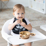 A smiling child wearing a black bib in a high chair eats fruit with Bumkins Silicone First Feeding Set: Minnie Mouse Gold in a bright room.