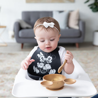 A baby in a high chair with a bow wears a black Mickey & Minnie bib as they eat from Bumkins Silicone First Feeding Set: Minnie Mouse Gold.