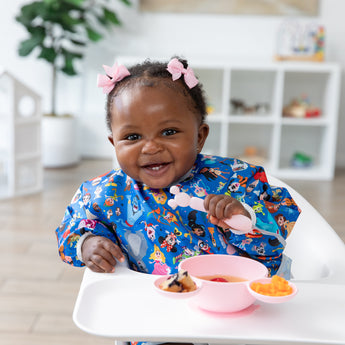 A baby in a high chair wearing a bib eats from a Bumkins Silicone First Feeding Set: Minnie Mouse Light Pink with a plant in the background.