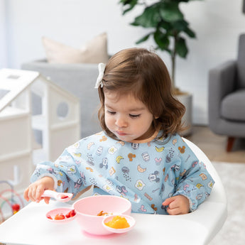 A girl wearing a blue bib in a high chair explores baby led weaning with Bumkins Silicone First Feeding Set: Minnie Mouse Light Pink.