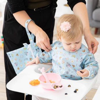 A child sits in a high chair with a Bumkins Silicone First Feeding Set: Minnie Mouse Light Pink on the tray; an adult behind adjusts their bib.