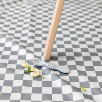 A Bumkins Silicone Dipping Spoons 3 Pack: Taffy lays atop a gray & white checkered mat among pieces of pasta & broccoli by a high chair leg.