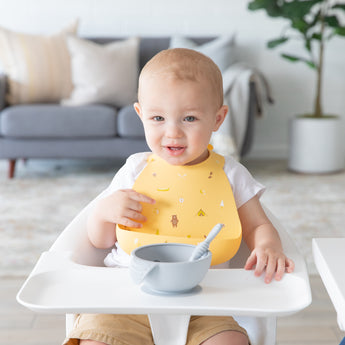 A baby wearing a yellow Bumkins Silicone Bib 2 Pack: Ocean Life and Camp Gear sits in a high chair with a gray bowl & spoon on the tray.