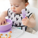 Close-up of a baby eating messily from a purple bowl while holding a Bumkins Silicone Chewtensils®: Purple Jelly.