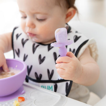 A baby holds a utensil from Bumkins First Foods Essential 5-Piece Mealtime Set: Purple Jelly while eating from a purple bowl.