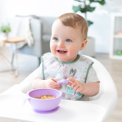 A baby with light brown hair sits in a high chair, wearing a bib and holding a spoon. The baby looks happy and has a small bowl of cereal in front of them. The background features a living room with a plant and furniture.