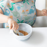 Close-up of a child in a dinosaur print bib eating applesauce from a bowl with a Bumkins Silicone Dipping Spoons 3 Pack: Taffy.