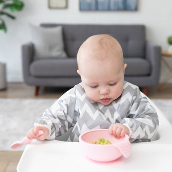 A baby in a gray & white bib eats mashed avocado with Bumkins Silicone First Feeding Set: Pink while seated in a high chair.