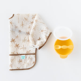 Bumkins Silicone First Feeding Set: Pineapple rests next to a folded beige & brown sleeved bib on a white background.