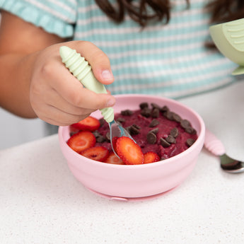 Close-up of a child using a green-handled spoon to place berries in a Bumkins Silicone Grip Bowl: Pink filled with acai & chocolate chips.