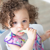 A curly haired girl in a floral bib places a Bumkins Silicone Dipping Spoons 3 Pack: Taffy in her mouth.