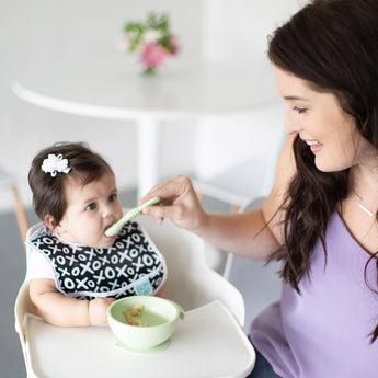 A woman feeds a baby wearing a bib in a high chair using a bowl and spoon from Bumkins Baby-Led Weaning Essential 6-Piece Set: Sage.
