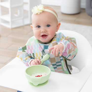 A smiling child sits in a high chair, wearing a flower headband and a colorful patterned bib while eating from a green bowl with a spoon.