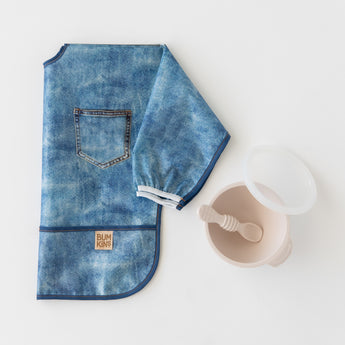 Bumkins Silicone First Feeding Set: Sand rests next to a folded denim print sleeved bib on a white background.