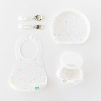 Bumkins Silicone First Feeding Set: Vanilla Sprinkle sits among a matching bib, plate, and utensils on a white background.