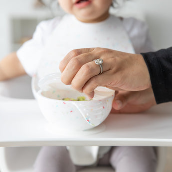An adult opens a Bumkins Silicone First Feeding Set: Vanilla Sprinkle as a toddler in a high chair watches, wearing a bib.