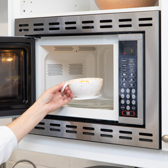 A person places a Bumkins Silicone First Feeding Set: Vanilla Sprinkle filled with food into an open microwave.