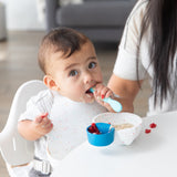 A baby sits in a high chair with an adult nearby, eating from a Bumkins Silicone First Feeding Set: Vanilla Sprinkle, wearing a matching bib.