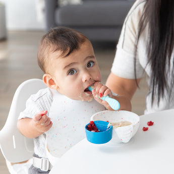A baby sits in a high chair with an adult nearby, eating from a Bumkins Silicone First Feeding Set: Vanilla Sprinkle, wearing a matching bib.