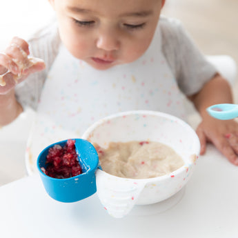 A toddler eagerly reaches into a Bumkins Silicone First Feeding Set: Vanilla Sprinkle filled with food, wearing a matching bib.