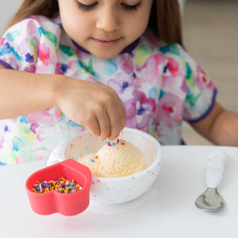 A child in a colorful bib adds rainbow sprinkles to ice cream in a Bumkins Silicone First Feeding Set: Vanilla Sprinkle at a white table.
