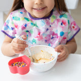 A child in a floral bib beams while enjoying ice cream from a Bumkins Silicone First Feeding Set: Vanilla Sprinkle  at a white table.