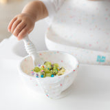 A child's hand grips a spoon, hovering over diced kiwi in a Bumkins Silicone First Feeding Set: Vanilla Sprinkle while wearing a matching bib.