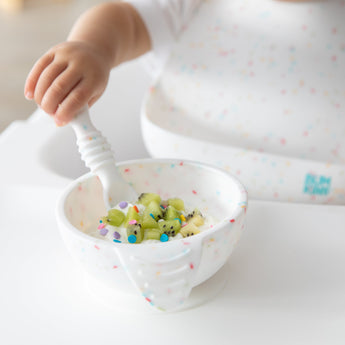A child's hand grips a spoon, hovering over diced kiwi in a Bumkins Silicone First Feeding Set: Vanilla Sprinkle while wearing a matching bib.
