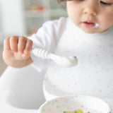 A toddler in a high chair holds a spoon with yogurt, eating from a Bumkins Silicone First Feeding Set: Vanilla Sprinkle.