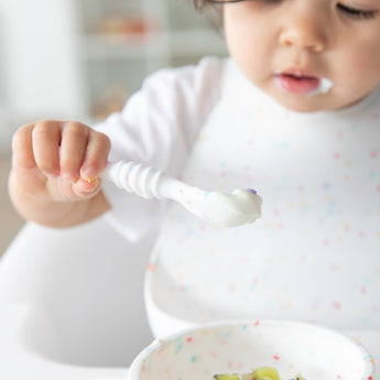 A toddler in a high chair holds a spoon with yogurt, eating from a Bumkins Silicone First Feeding Set: Vanilla Sprinkle.