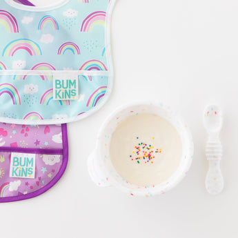 Two colorful bibs lay next to a Bumkins Silicone First Feeding Set: Vanilla Sprinkle filled with yogurt and sprinkles on a white background.