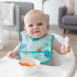 A baby wearing a Bumkins Starter Bib 2 Pack: Ocean Life & Whale Tail sits in a high chair, eating mashed sweet potatoes from a white bowl.
