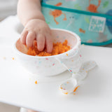 A baby's hand reaches into a Bumkins Silicone First Feeding Set: Vanilla Sprinkle filled with mashed sweet potato with a spoon beside it.