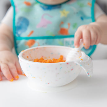 Close-up of a baby wearing a colorful bib enjoying orange food from a Bumkins Silicone First Feeding Set: Vanilla Sprinkle at a white table.