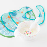 Bumkins Silicone First Feeding Set: Vanilla Sprinkle filled with yogurt & sprinkles rests in front of two ocean-themed waterproof bibs.