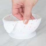 A hand holds a Bumkins Silicone First Feeding Set: Vanilla Sprinkle, demonstrating the suction cup bottom on a marble surface.