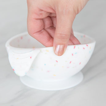 A hand holds a Bumkins Silicone First Feeding Set: Vanilla Sprinkle, demonstrating the suction cup bottom on a marble surface.