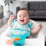 A smiley baby wearing a Bumkins Starter Bib 2 Pack: Ocean Life & Whale Tail sits in a high chair with a blue bowl of mashed food on the tray.