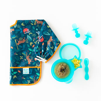 A folded Bumkins Sleeved Bib 2 Pack: Jungle and Animal Print lays next to a blue bowl with applesauce & blue utensils on a white background.