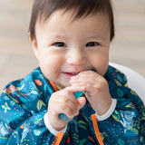 A smiling baby wearing a Bumkins Sleeved Bib 2 Pack: Jungle and Animal Print chews a blue spoon.
