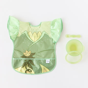 Bumkins Ruffle Apron Bib: Tiana lays next to a green bowl, spoon, & lid set on a white background.