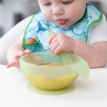 Close-up of a baby exploring mealtime with a bowl & spoon from Bumkins First Foods Essential 5-Piece Mealtime Set: Green Jelly.