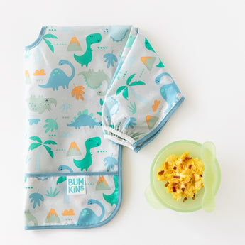 Bumkins Sleeved Bib: Dinosaurs lays folded on a white background next to a green bowl with eggs and a green spoon.