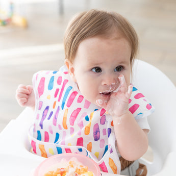 In a high chair, a baby with a colorful bib enjoys messy mealtime using Bumkins Silicone First Feeding Set: Pink Jelly.