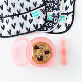 Bumkins Silicone First Feeding Set: Pink Jelly filled with oatmeal and berries on a white background, with black and white bibs nearby.