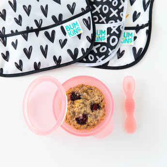 Bumkins Silicone First Feeding Set: Pink Jelly filled with oatmeal and berries on a white background, with black and white bibs nearby.