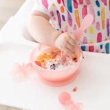 A baby in a colorful bib grips a spoon and a Bumkins Silicone First Feeding Set: Pink Jelly filled with yogurt and fruit on a high chair.