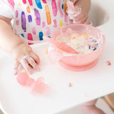 Close-up of a child in a high chair reaching for a Bumkins Silicone Chewtensils®: Pink Jelly while messily eating yogurt and fruit from a bowl.