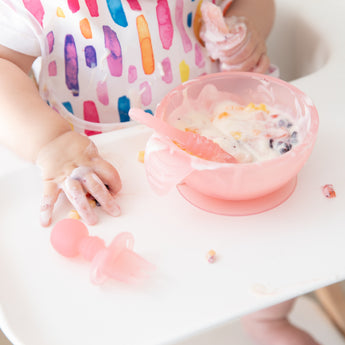Close-up of a child in a high chair reaching for a Bumkins Silicone Chewtensils®: Pink Jelly while messily eating yogurt and fruit from a bowl.