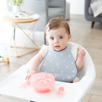 A girl wearing a gray bib in a high chair eats yogurt with Bumkins First Foods Deluxe 6-Piece Mealtime Set: Pink Jelly.